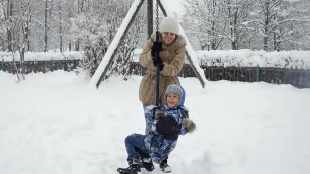 雪の冬の日に遊び場で遊んでジップラインに乗っている彼女の息子と幸せな母親 冬の休暇 一緒に遊ぶ家族 雪の中での活動の楽しさと喜び — ストック動画