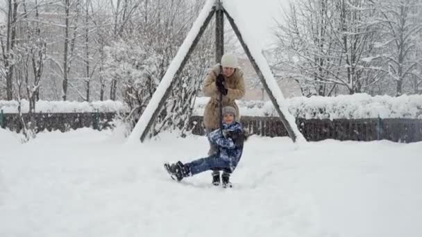 雪に覆われた公園でジップラインで彼女の息子に乗って幸せな陽気な母親のスローモーション 冬の休暇 一緒に遊ぶ家族 雪の中での活動の楽しさと喜び — ストック動画