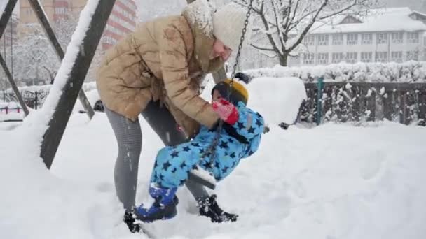 吹雪の後 雪に覆われた遊び場でスイングに彼女の赤ちゃんの息子と幸せな母親 冬休みの楽しみと喜び 屋外で遊ぶ子供たち 雪の中での活動 — ストック動画