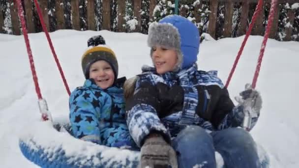 吹雪の後に雪に覆われた公共の遊び場でスイングを楽しんでいる弟と一緒に幸せな笑顔の兄 冬休みの楽しみと喜び 屋外で遊ぶ子供たち 雪の中での活動 — ストック動画
