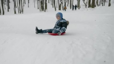 Yüzünde kocaman bir gülümsemeyle kar kaplı bir tepede kayarak trambolinde zıplayan bir çocuğun yavaş çekim görüntüleri. Noel tatili boyunca çocuklar için mükemmel bir kış aktivitesi.