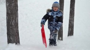 Kar yağarken plastik kızaklarıyla karlı tepeye tırmanan mutlu çocuk. Çocukların kış boyunca eğlenmesi, Noel tatilleri ve dışarıda karda oynaması.