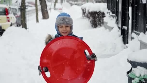 冬の公園の雪でプラスチック製のそりで歩く陽気な少年のスローモーション クリスマス休暇 雪の中で屋外で遊ぶ子供たちの概念 — ストック動画