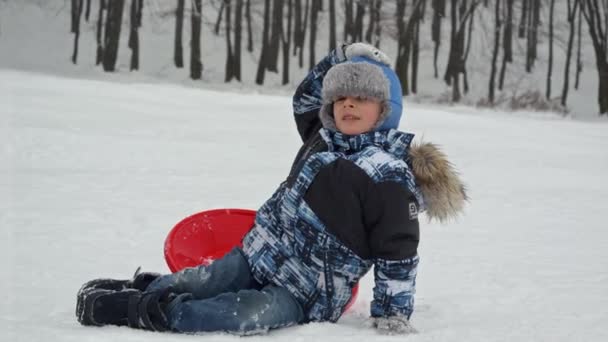 動揺した少年はそりから落ち 雪の丘を滑り降りると頭を痛めた 活動中の冬の休日や極端なスポーツでの怪我や外傷 — ストック動画