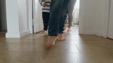 Evdeki ahşap zeminde koşan iki çocuk ve annesinin ayaklarının yakınlaşması. Aile mutluluğu kavramı, ebeveynlerin çocuklarla oynaması, evde eğlenmesi, neşe ve sevgi.