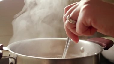 Kadın elinin kaşıkla metal tavada kaynamış çorbayı karıştırdığı yavaş çekim. Buhar yavaşça sıcak sudan yükseliyor.