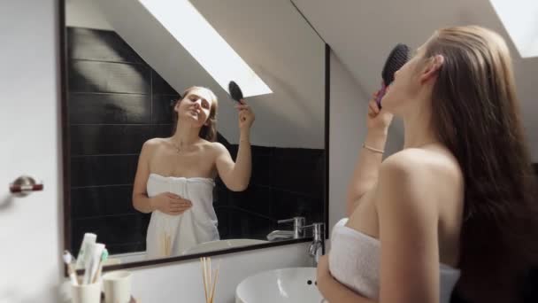 女人站在浴池里 披着毛巾 一边梳理长发 一边看着镜子里的自己 保持良好卫生习惯和自我照顾的概念 — 图库视频影像