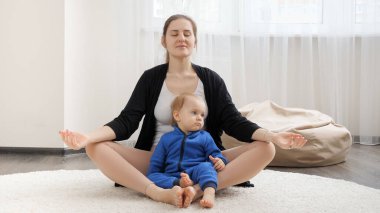 Nilüfer pozisyonunda meditasyon yapan genç ve güzel bir anne ve 1 yaşındaki bebeği evde. Aile sağlığı, aktif yaşam tarzı, ebeveynlik ve çocuk gelişimi.