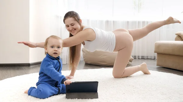 幸せな笑顔母とともに赤ちゃん息子行うフィットネス運動とオンライントレーニングを見ますタブレットコンピュータ — ストック写真