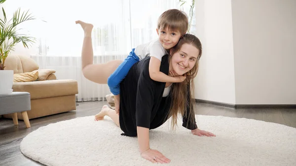 小男孩骑在妈妈的背上 而她正在家里做健身锻炼 家庭保健 积极的生活方式 养育子女和儿童发展 — 图库照片
