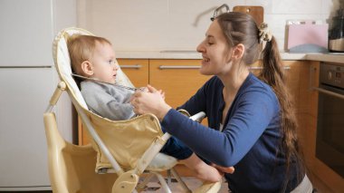 Genç anne, oğluna yemek vermeden önce bebek sandalyesine emniyet kemeri takıyor. Ebeveynlik kavramı, sağlıklı beslenme ve bebek bakımı