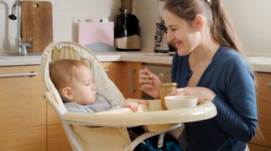 Young mother smiling at her baby son sitting in highchair at kitchen. Concept of parenting, healthy nutrition and baby care