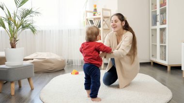 Mutlu kahkahalar atan anne, bebeğinin ona doğru yürümesini destekliyor ve tezahürat yapıyor. Bebek gelişimi, aile oyunları, ilk adımlar, ebeveynlik ve bakım.