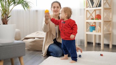 Küçük çocuk annesinden renkli oyuncaklar alıyor ve oturma odasında yumuşak halıda yürüyor. Bebek gelişimi, aile oyunları, ilk adımlar, ebeveynlik ve bakım.