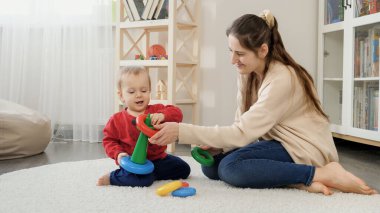 Annesiyle birlikte küçük bebek renkli oyuncak piramidini oturma odasında yerde monte etmeyi öğreniyor. Bebek gelişimi, çocuk oyunları, eğitim ve öğrenim