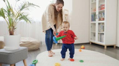 Bebek gibi gülümseyen mutlu çocuk, elinde oyuncaklar, oturma odasında halıda yürümeyi öğreniyor. Bebek gelişimi, aile oyunları, ilk adımlar, ebeveynlik ve bakım.