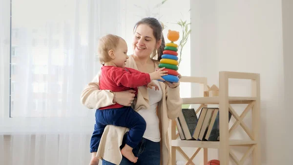居間の棚から赤ん坊の息子におもちゃを与える幸せな笑顔の母 赤ちゃんの発達 家族で遊ぶゲーム 第一歩を踏み出す 親とケア — ストック写真