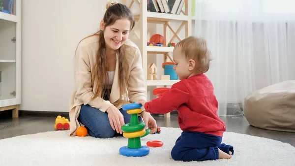 Happy smiling mother looking at her little baby son assembling toy pyramid or tower. Baby development, child playing games, education and learning