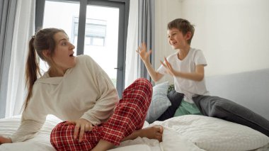 Küçük çocuk yastık yığınının altından atlıyor ve annesini yatakta otururken korkutuyor. Mutlu aile kavramı, çocuklu ebeveynler, olumlu duygular ve evde rahatlama.