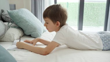 Küçük çocuk pijama giyip sabah uyandıktan sonra yatakta ödev yapıyor. Evde eğitim, çocuk gelişimi ve uzaktan eğitim kavramı