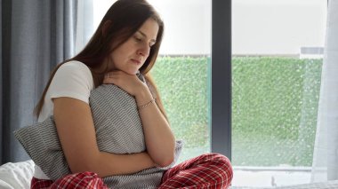 Pijamalı, düşünceli genç bir kadın yatakta oturup mindere sıkıca sarılıyor. Depresyon, olumsuz duygular, yalnızlık ve stres kavramı