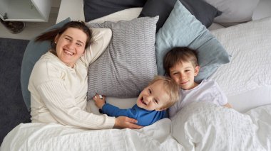 Mutlu bir anne ve iki oğlu sabah yatakta yatıp kameraya gülümsüyor. Aile mutluluğu kavramı, evde dinlenmek, yatakta eğlenmek, ebeveynler ve neşeli çocuklar..