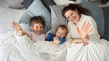 Genç mutlu anne iki oğluyla yatakta uzanıyor ve kameraya el sallıyor. Aile mutluluğu kavramı, evde dinlenmek, yatakta eğlenmek, ebeveynler ve neşeli çocuklar.