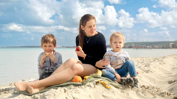 Młoda Kobieta Dwoma Synami Organizującymi Piknik Piaszczystej Plaży Morskiej Jedzącymi — Zdjęcie stockowe