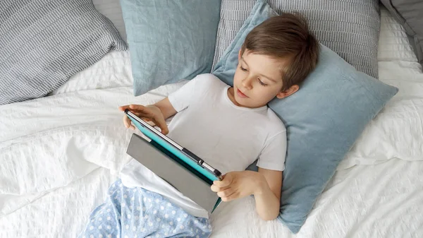 小さな男の子がベッドの上に寝そべっていて 昼間はタブレットコンピュータを使っている ガジェット 教育と開発を使用して子供 — ストック写真