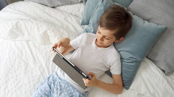 昼間はベッドの上に寝そべっている間 タブレットPcで遊んでいると 男の子は喜んで笑っています ガジェット 教育と開発を使用して子供 — ストック写真