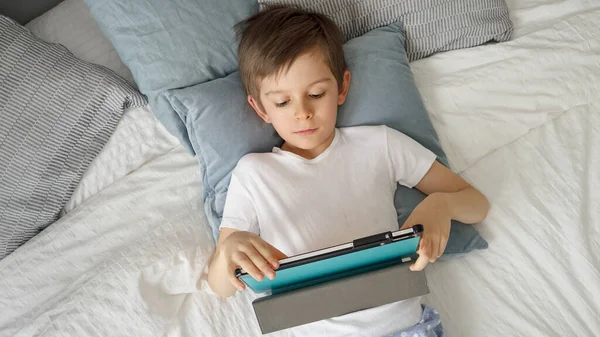 Szczęśliwy Młody Chłopiec Bawi Się Tabletem Leżąc Łóżku Ciągu Dnia — Zdjęcie stockowe