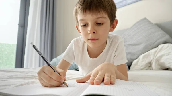 小男孩躺在床上 一边做作业 一边在笔记本上写字 儿童发展和在家远程学习 — 图库照片