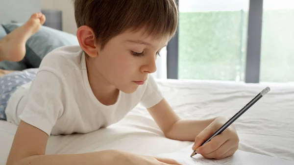 Mały Chłopiec Odrabia Pracę Domową Łóżku Pisze Notatniku Pilnością Poświęceniem — Zdjęcie stockowe