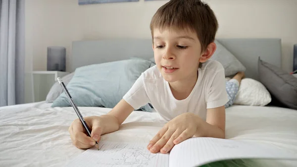 ベッドに寝そべっている間にノートに書いたパジャマ姿の少年が宿題をしている 遠隔学習環境における教育と子どもの発達 — ストック写真