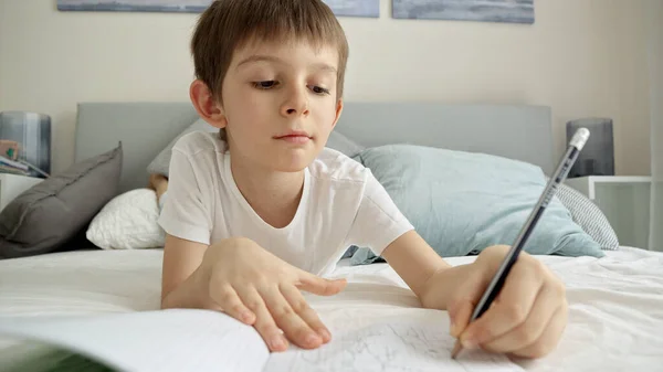 小さな男の子がベッドで宿題をしてノートを書いている 家庭からの教育 子供の発達 遠隔教育の概念 — ストック写真