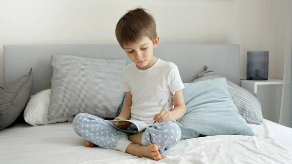 小さな男の子がベッドに座って鉛筆でノートに書きます 家庭での教育 子供の発達 遠隔教育の概念 — ストック写真