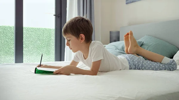 大きな窓のベッドの上に寝そべってノートに宿題をしているパジャマ姿の少年 家庭での教育 子供の発達 遠隔教育の概念 — ストック写真