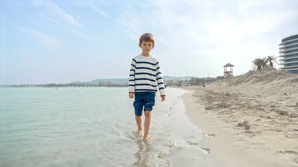 Cute Uśmiechnięty Chłopiec Swetrze Pluskając Wodą Morską Jak Chodzi Piaszczystej — Zdjęcie stockowe