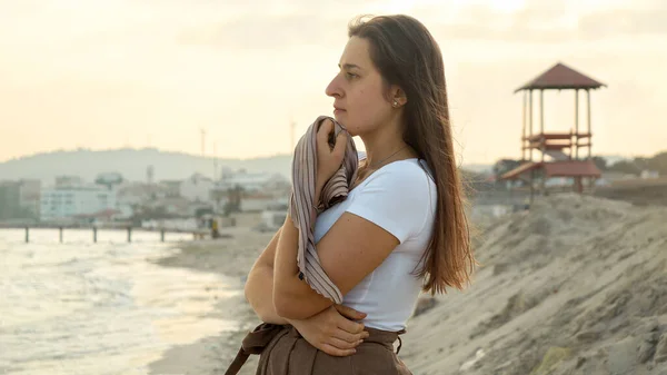 砂浜に立ち 波に揺れるシルクのスカーフを持ち 日没時に海を眺める真剣な表情の穏やかな若い女性 — ストック写真