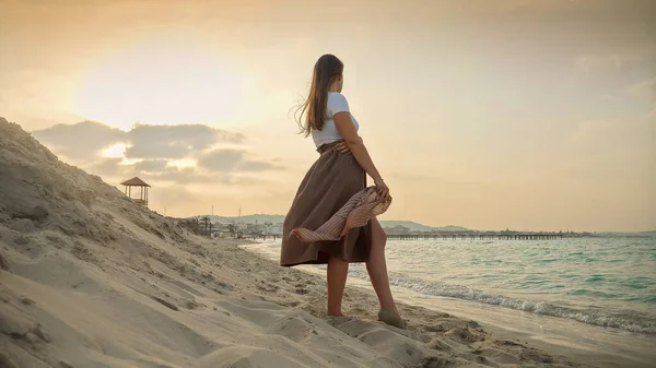 平静的年轻女子站在沙滩上 拿着一条飘扬的丝巾 凝视着海面上的夕阳 — 图库照片