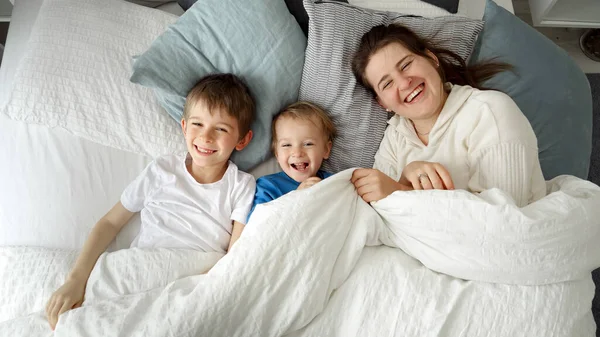Szczęśliwa Matka Synami Leżącymi Łóżku Pod Kołdrą Uśmiechającymi Się Kamery — Zdjęcie stockowe