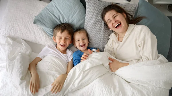 カメラに手を振っている2人の息子と一緒にベッドの中で明るい若い母親 これは 家族の幸福の本質を捉え 家庭で質の高い時間を過ごし 親子の楽しい瞬間を捉えます — ストック写真
