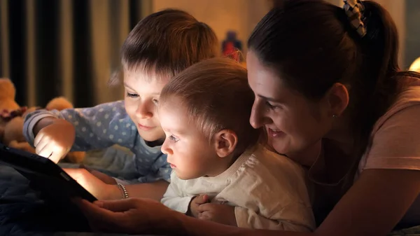 夜にベッドの上で母親とテーブルコンピュータでゲームをプレイする2人の幸せな男の子の肖像画 一緒に時間を過ごす家族 子育て 幸せな子供時代とエンターテイメント — ストック写真