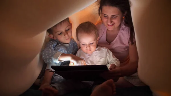 ベッドシートの下に隠れてタブレットPcでゲームをしている母親を持つ2人の男の子 一緒に時間を過ごす家族 子育て 幸せな子供時代とエンターテイメント — ストック写真