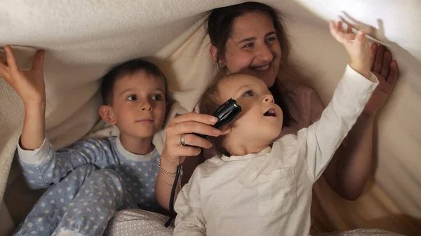 夜に毛布の下に隠れている間 懐中電灯で遊んでいるパジャマ姿の2人の男の子と母親 一緒に時間を過ごす家族 子育て 幸せな子供時代とエンターテイメント — ストック写真