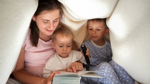 2人の息子の夜に毛布の下に懐中電灯でおとぎ話の本を読んで若い母親 一緒に時間を過ごす家族 子育て 幸せな子供時代とエンターテイメント — ストック写真