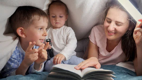 一个笑着的女人 两个儿子躺在毯子下 看睡前故事书 有时间在一起的家庭 养育子女 快乐的童年和娱乐 — 图库照片
