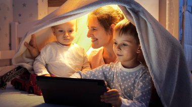 Yatakta battaniye çadırında oturan ve tablet bilgisayar kullanan iki çocukla gülümseyen anne. Ailenin birlikte vakit geçirmesi, aletli çocuklar, ebeveynlik, mutlu bir çocukluk ve eğlence.