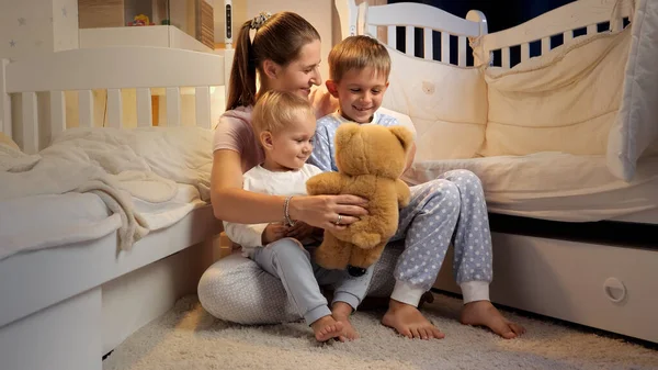 傍晚时分 年轻可爱的妈妈穿着睡衣和泰迪熊在孩子们的房间里和两个儿子玩耍 有时间在一起的家庭 养育子女 快乐的童年和娱乐 — 图库照片