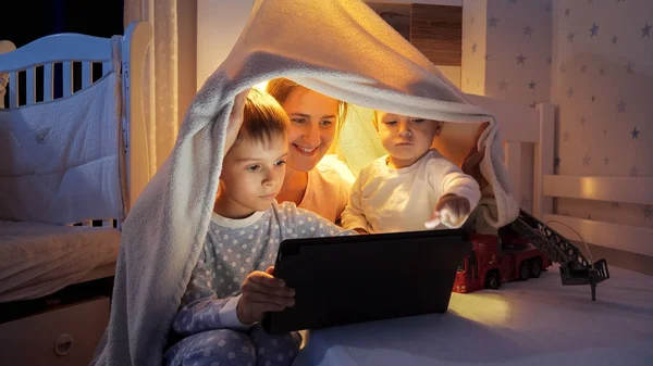 快乐的一家人晚上在卧室里用平板电脑看卡通片 有时间在一起的家庭 带着小玩艺儿的孩子 快乐的童年和娱乐 — 图库照片